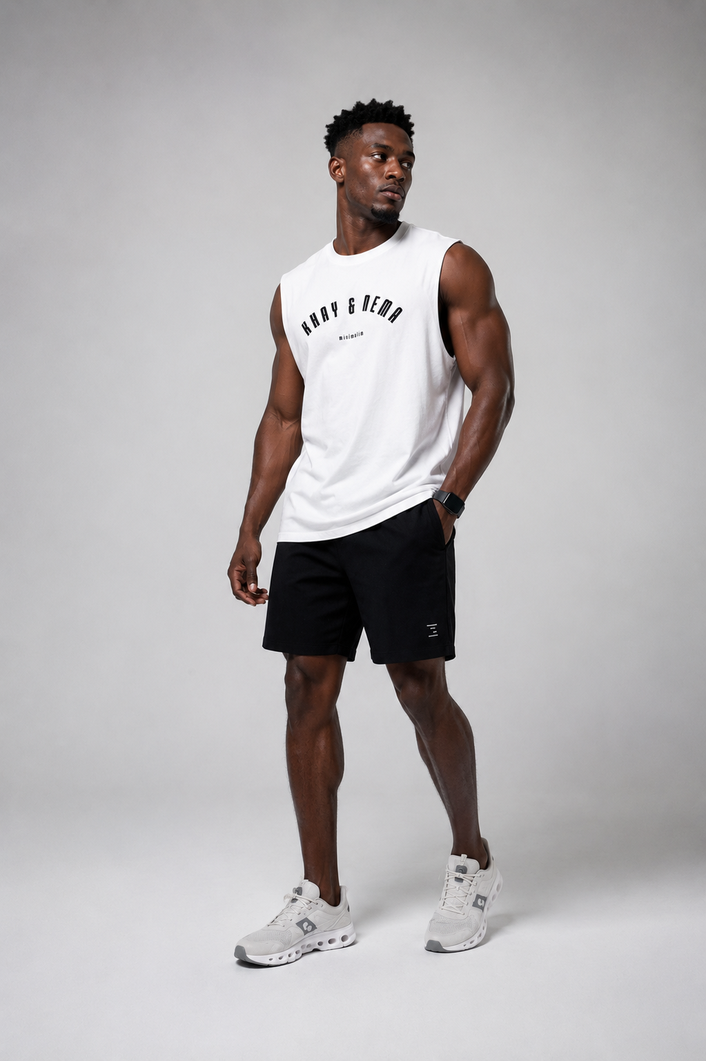 A man modeling a white sleeveless athletic tank top with black text and black athletic shorts, paired with white sneakers, against a light gray studio background.