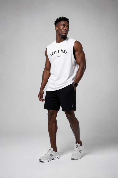 A man modeling a white sleeveless athletic tank top with black text and black athletic shorts, paired with white sneakers, against a light gray studio background.